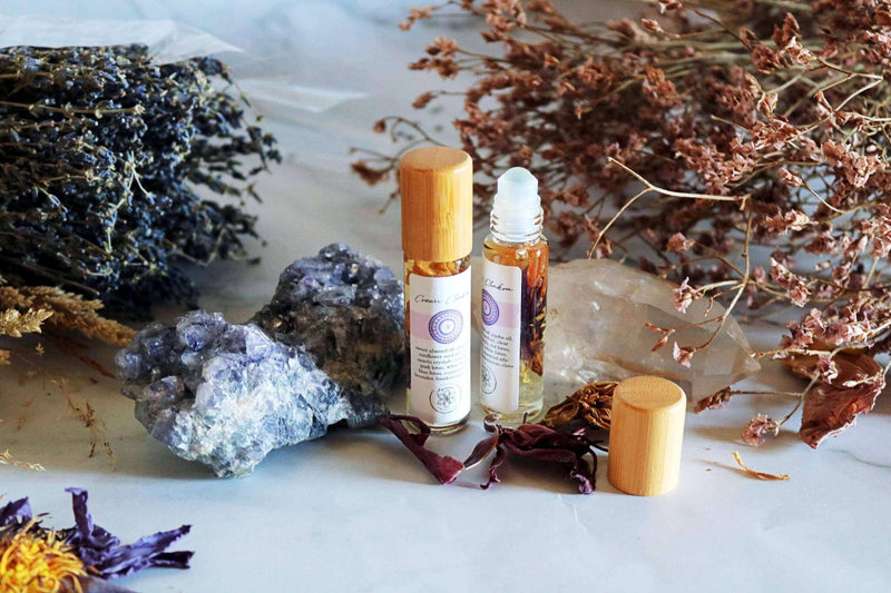 Zwei Roll-on-Flaschen mit Chakra-Öl und Holzdeckeln stehen zwischen getrocknetem Lavendel, braunen Blumen, einem großen blauen Fluoritkristall und einem Quarzkristall auf weißem Hintergrund.