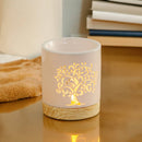 Eine beleuchtete, weiße Keramik-Duftlampe mit leuchtendem Lebensbaum-Ausschnitt und Holzsockel, auf einer reflektierenden Oberfläche neben einem Stapel Bücher.