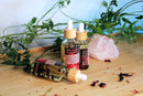 Drei Tropfflaschen "Hot n Spicy Tantric Massage Oil" sind auf einem hellen Holztablett arrangiert. Eine Flasche liegt auf der Seite und zeigt rote Hibiskusblütenblätter im klaren Öl, während die anderen beiden aufrecht stehen. Ein roher rosa Rosenquarzkristall und grüne belaubte Zweige schmücken den Hintergrund vor einer hellblauen Wand.