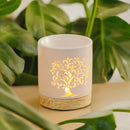 Eine beleuchtete, weiße Keramik-Duftlampe mit leuchtendem Lebensbaum-Ausschnitt und Holzsockel, vor einem Hintergrund aus üppig grünen tropischen Blättern.