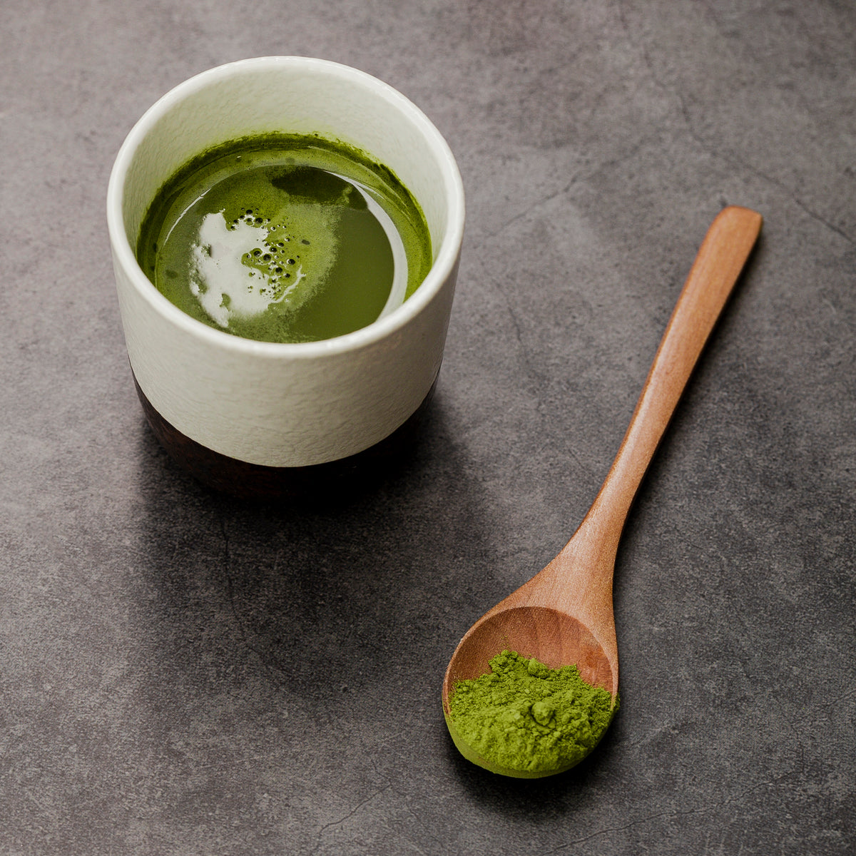 Eine Tasse Matcha-Tee mit grünem Pulver in einem Holzlöffel auf dunklem Steinuntergrund.