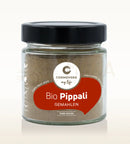 Ein durchsichtiges glas mit schwarzem deckel enthält cosmoveda pippali gemahlen bio pippali für ayurvedische küche