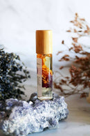 Eine Roll-on-Flasche mit Chakra-Öl und einem Holzdeckel steht auf einem leuchtend blauen und lila Fluoritkristall. Im Hintergrund sind getrockneter Lavendel und braune Pflanzen zu sehen.