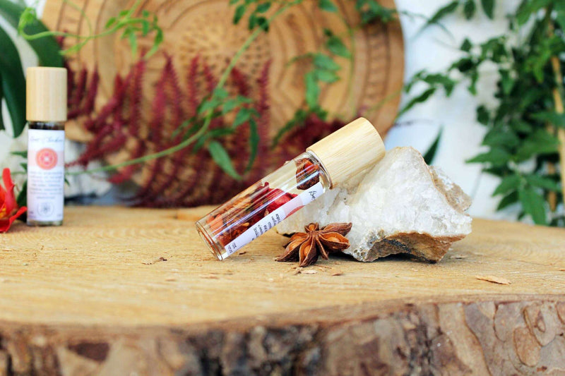 Auf einer naturbelassenen Holzscheibe liegt eine Roll-On-Flasche mit Bambusverschluss, gefüllt mit roten Botanika, horizontal an einem großen, klaren Kristall. Eine Sternanis-Kapsel liegt vor der Flasche, und im unscharfen Hintergrund sind eine zweite, aufrechte Roll-On-Flasche sowie üppiges grünes und tiefrotes Laub sichtbar.