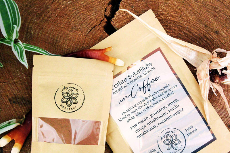 Zwei „Kaffee-Ersatz“-Superfood-Pulvermischungen von „Ayamanu Naturals“ sind kunstvoll auf einer strukturierten Holzoberfläche arrangiert. Ein Beutel zeigt seinen Inhalt, während der andere ein detailliertes Etikett mit Zutaten aufweist. Natürliche Elemente wie ein auffälliges rot-weiß gesprenkeltes Blatt und getrocknete Maiskolben verstärken die organische Präsentation.