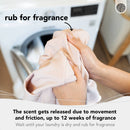 Eine Nahaufnahme, die zeigt, wie die Hände einer Person ein Stück trockene, helle Wäsche reiben, im Hintergrund unscharf eine Waschmaschine. Der Text besagt „rub for fragrance“ (reiben für Duft) und erklärt: „Der Duft wird durch Bewegung und Reibung freigesetzt, bis zu 12 Wochen Duft / Warten Sie, bis Ihre Wäsche trocken ist, und reiben Sie für Duft.“