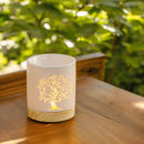 Eine beleuchtete, weiße Keramik-Duftlampe mit leuchtendem Lebensbaum-Ausschnitt, die draußen auf einer Holzoberfläche mit einem üppig grünen Hintergrund steht.