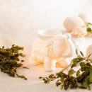 Beleuchtete weiße Keramik-Duftlampe in Elefantenform, umgeben von Pflanzen auf einem hellen Tuch.