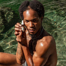 Ein Mann mit dunklen, lockigen Haaren und einer Kette sitzt in klarem grünen Wasser und hält eine kleine Pipettenflasche mit Gesichtsserum hoch.