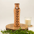 Eine glänzende, gehämmerte Kupfer-Trinkflasche mit Wabenmuster, die auf einem runden Holzpodest neben einer brennenden Kerze steht.