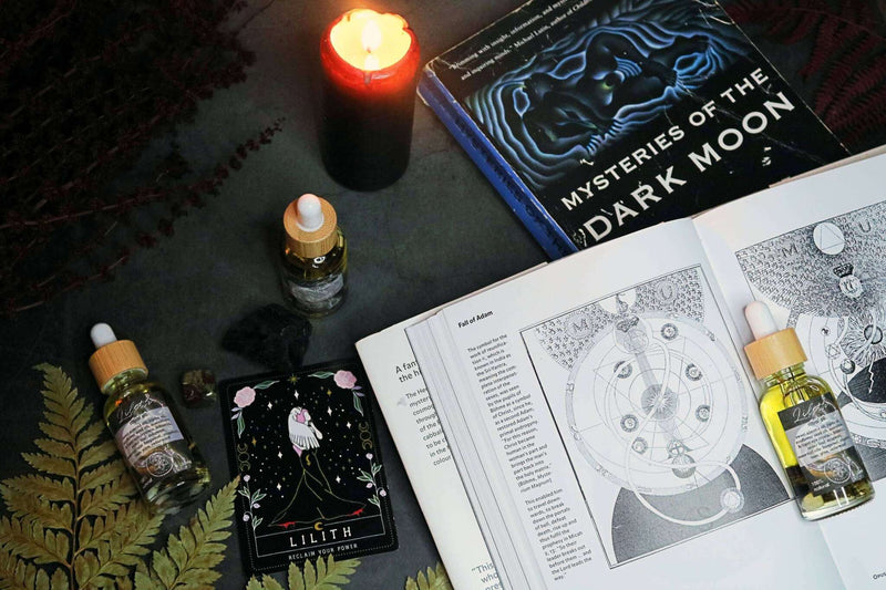 Eine mystische Anordnung auf einer dunklen Oberfläche zeigt eine brennende schwarze Kerze, eine aufrechte Ritualöl-Flasche mit Pipette, einen schwarzen Rohkristall, eine "Lilith"-Orakelkarte und ein offenes Buch. Ein dunkles Buch "Mysteries of the Dark Moon" ist ebenfalls im Hintergrund sichtbar, zusammen mit getrocknetem grünem und rotem Laub.