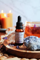 Eine Flasche "Blue Lotus Tinktur" steht auf einer Gruppe violetter Kristalle auf einem Holztablett. Im Hintergrund sind verschwommen zwei brennende Kerzen, ein Glas bernsteinfarbener Tee mit einer violetten Blume und klare Quarzkristalle zu sehen, die eine ruhige, warme Atmosphäre schaffen