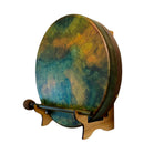 Eine runde Handtrommel (Rahmen- oder Schamanentrommel) mit einem bemalten Fell in lebhaften, gesprenkelten Blau-, Grün-, Gelb- und Orangetönen, schräg auf einem kleinen Holzständer stehend, mit einem hölzernen Schlägel davor platziert.