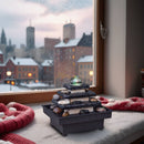Ein mehrstufiger Zen-Zimmerbrunnen aus Stein mit leuchtender Glaskugel steht auf einem verschneiten Fensterbrett vor einer gemütlichen, winterlichen Stadtlandschaft in der Dämmerung.