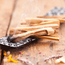 Ein glimmendes Palo-Santo-Holzstäbchen, das dünnen Rauch auf einer gemusterten schwarzen Schale über einer Holzoberfläche abgibt.