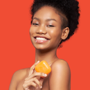 Eine lächelnde Frau hält ein orangefarbenes Stück Seife vor einem leuchtend orangefarbenen Hintergrund. Ihr Gesichtsausdruck ist fröhlich und vermittelt ein Gefühl von Frische und Vitalität.