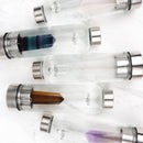 Eine Draufsicht auf fünf Glaswasserflaschen, jede mit einem andersfarbigen Kristall, diagonal auf einer weißen Marmorfläche angeordnet.