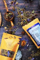 Eine Mischung aus losen Kräutern und getrockneten Blütenblättern ist auf einer dunklen Holzoberfläche verstreut, zusammen mit zwei Teepackungen, einem hölzernen Schöpflöffel und Kristallen.