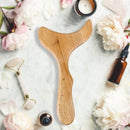 Hölzernes Gua-Sha-Massagegerät inmitten von Pflegeprodukten und rosa Pfingstrosen auf Marmor.