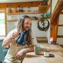 A man with long wavy hair and a beard stirs a steaming mug on a wooden table. Beside it, an open jar of Paavani Ashwagandha is visible.