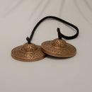 Ein Paar kleiner, verzierter Tingsha-Zimbeln aus Messing oder Bronze, verbunden durch eine schwarze Lederschnur, auf weißem Hintergrund.