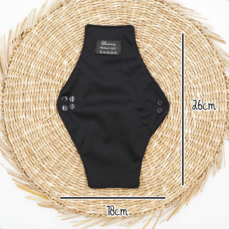 Eine schwarze wiederverwendbare Binde von Bamboozy (Medium Light) auf einer gewebten Matte, mit Maßen von 26cm Länge und 18cm Breite, ausgestattet mit Druckknöpfen