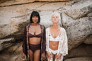 Zwei Frauen, eine mit dunklem Haar in braunem Bikini und gemustertem Morgenmantel, die andere mit platinblondem Haar in weißem Bikini und Spitzen-Überwurf, stehen vor großen Felsen. Die blonde Frau spitzt spielerisch die Lippen.