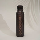 Dunkle, kupferfarbene Isolier-Wasserflasche, in deren Oberfläche die sieben Chakra-Symbole geätzt sind, vor einem schlichten Hintergrund mit Palmblattschatten.