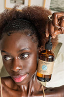  Eine Frau mit lockigem braunem Haar hält eine PAAVANI Pitta Körperölflasche mit hellblauem geometrischem Muster nahe ihrem Gesicht. Sie hat helle Augen und glänzende Lippen und blickt leicht nach rechts.