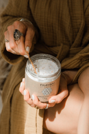 Eine Frau mit einem Ring und hell lackierten Nägeln schöpft mit einem kleinen Holzlöffel weißes Pulver aus einem Glas "PAAVANI AYURVEDA COCONUT MILK BATH bathing ritual" (240 g). Sie trägt einen locker sitzenden braunen Bademantel.