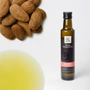 Eine Flasche Mandelöl von Ölwerk steht rechts auf einer weißen Oberfläche, mit einem Haufen ganzer Mandeln in der oberen linken Ecke und einem verschwommenen Tropfen des goldgelben Öls im unteren linken Bereich. Das Etikett der Flasche weist auf "feinste biologische Öle" und "100% rein & naturbelassen" hin.