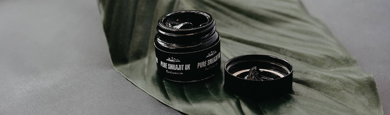 Ein geöffnetes Glas mit "PURE SHILAJIT UK Himalayan resin" auf einem großen grünen Blatt, zeigt die schwarze, teerartige Substanz und den Deckel daneben.
