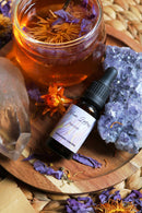 Aufnahme von oben von einer kleinen Flasche „Blue Lotus Tincture“ auf einem Holztablett neben einem großen Stück rohem blauem Amethyst und einem klaren Glasgefäß mit bernsteinfarbenem Tee, der getrocknete blaue Lotusblütenblätter und Staubgefäße enthält.