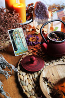 Eine Teekanne aus rotem Gusseisen mit Kräutern steht auf einem Tisch, umgeben von brennenden Kerzen, einem Tarotkarte, getrockneten Kräutern und Kristallen.