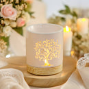 Eine beleuchtete, weiße Keramik-Duftlampe mit leuchtendem Lebensbaum-Ausschnitt, auf einem hölzernen Untersetzer inmitten weicher Stoffe, Blumen und brennender Kerzen.