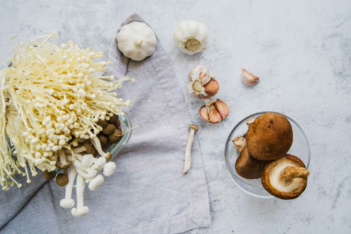 Frische Enoki-Pilze, Knoblauchzehen und Shiitake-Pilze in Schalen auf hellem Untergrund.