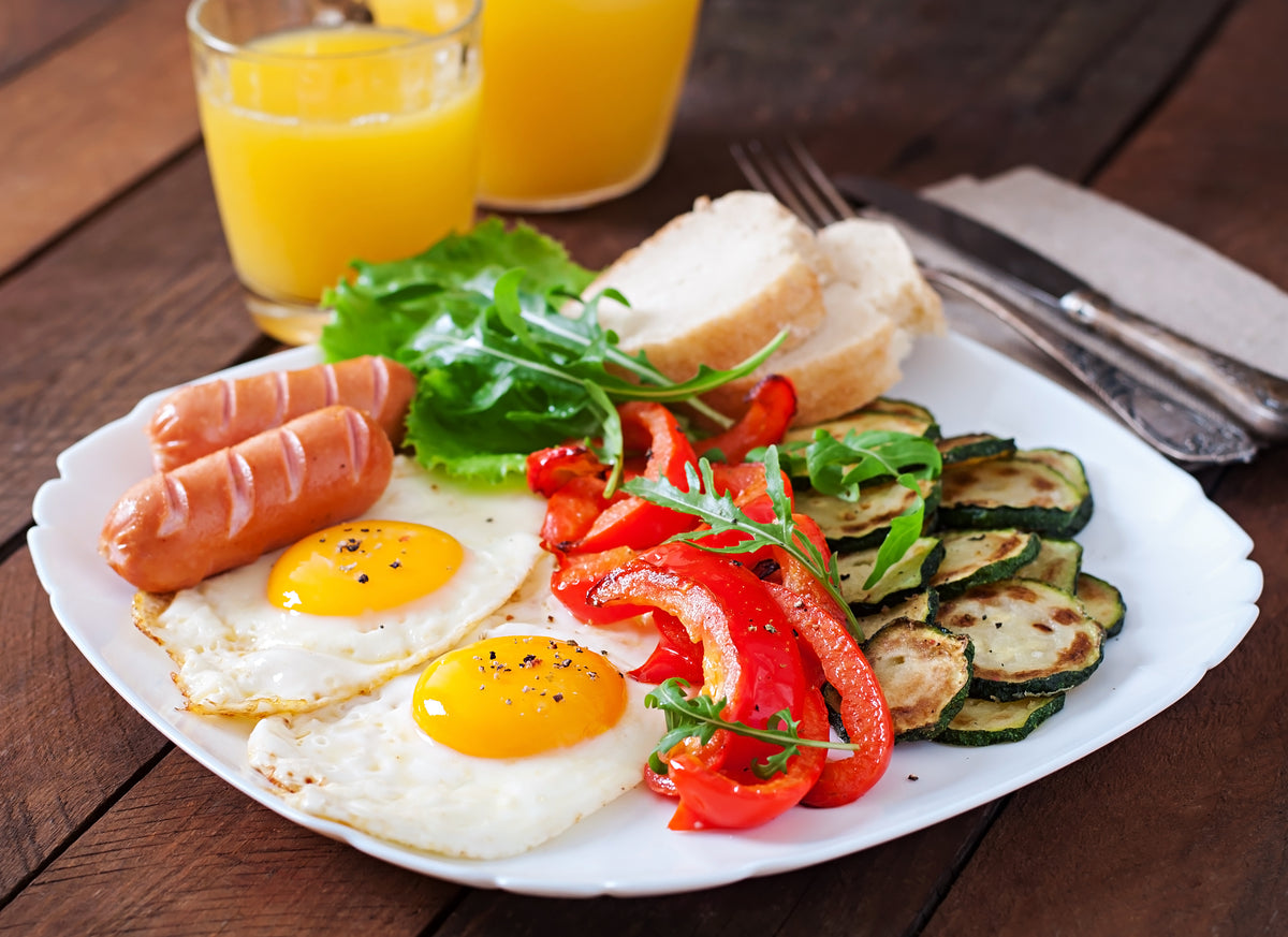 Frühstücksteller mit Spiegelei, Würstchen, gebratenem Gemüse und frischem Orangensaft.