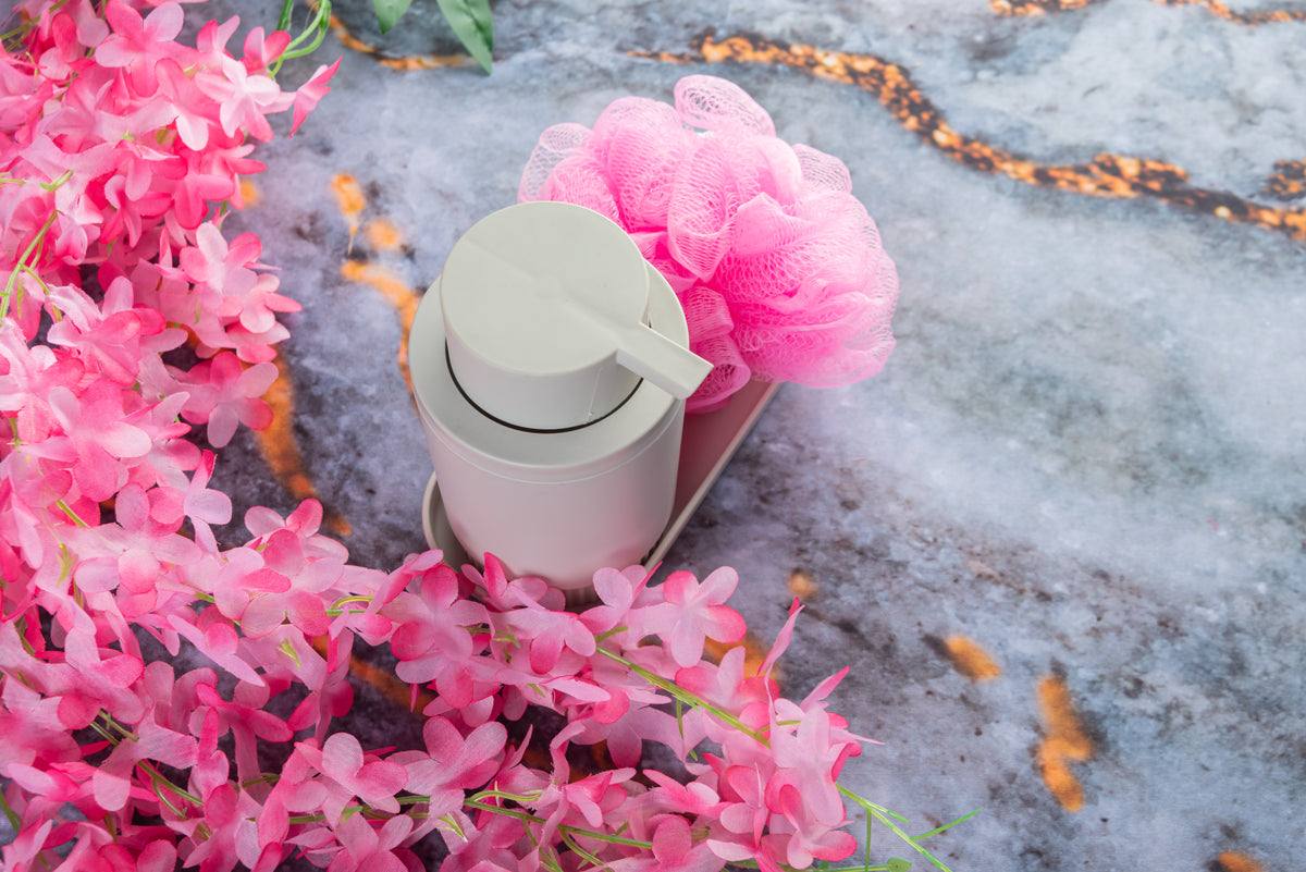 Weiße Seifenspender-Flasche mit rosa Duschschwamm und dekorativen rosa Blüten auf Marmor.