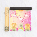 Eine teilweise ausgerollte Myga Yogamatte mit einem leuchtenden, tropischen Design aus pinkfarbenen Flamingos und großen gelben Ananas auf einem bunten Hintergrund, neben ihrer braunen Kartontragrolle.