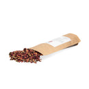 Getrocknete Rosenblüten fallen aus einem offenen braunen Papierbeutel auf eine weiße Oberfläche.