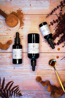 Drei dunkle Pipettenflaschen mit Reishi-Flüssigextrakt auf einer Holzoberfläche mit pflanzlichen Inhaltsstoffen.