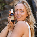 Eine blonde Frau lächelt und hält eine Flasche Paavani Ayurveda Vata Toner für trockene Haut hoch.