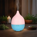Ein rosa-blauer tropfenförmiger Aroma-Diffusor, der Nebel ausstößt und sanft beleuchtet auf einem Holztisch mit Kerze und Kräutern im Hintergrund steht.