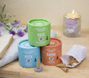 Drei bunte Behälter mit ayurvedischen Räucherkegeln neben einer brennenden Kerze und Blumen.