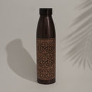 Dunkle, kupferfarbene Isolierflasche mit verziertem, geätztem Muster, vor einer weißen Wand mit dem Schatten eines Palmblattes.