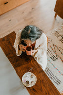 Eine Aufnahme von oben zeigt eine Frau mit grau meliertem Haar in einem weißen Bademantel, die elegant aus einer pinkfarbenen Tasse trinkt. Auf dem Holztisch sind ein Teller mit verschütteten Teeblättern, ein Löffel und eine kleine Teedose aus Metall zu sehen.