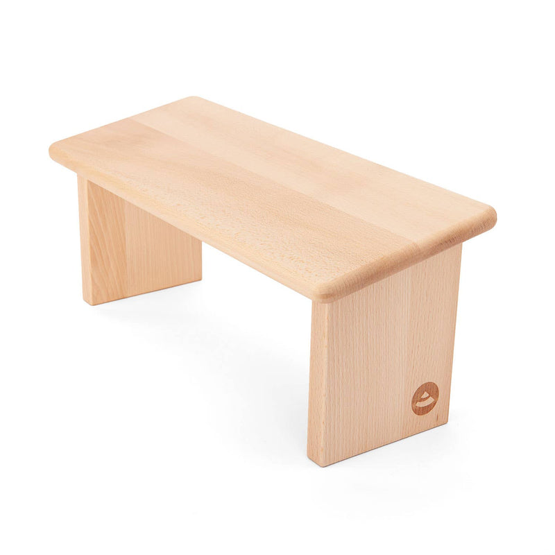 Helle Buchenholz-Meditationsbank mit glatter rechteckiger Sitzfläche und zwei massiven Beinen, dezente Logo-Gravur an einem Bein, auf weißem Hintergrund.