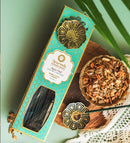 Eine hohe, türkis-goldene Schachtel Ayurveda Aqua Oud Räucherstäbchen (Kühlend + Beruhigend - Pitta) mit einem messingfarbenen, blumenförmigen Medaillon oben. Sie steht auf einer Holzoberfläche neben einer kleinen Messingschale mit Holzspänen und einem kleineren Räucherstäbchenhalter.