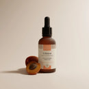 Eine gefrostete Glas-Tropfflasche mit „L'Abricot“ Illuminating-Öl von Comptoir des Huiles neben einer frischen halbierten Aprikose.