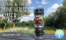 Ein Werbebild mit einem hohen, gestapelten Glas, gefüllt mit Schichten von Brombeeren, Erdbeeren und Blaubeeren, das auf einem Tisch im Freien neben einem See steht. Der Text lautet: "ENERGISE & THRIVE – DRINK HEALTHY WATER" (Energetisieren & Gedeihen – Trinken Sie gesundes Wasser).